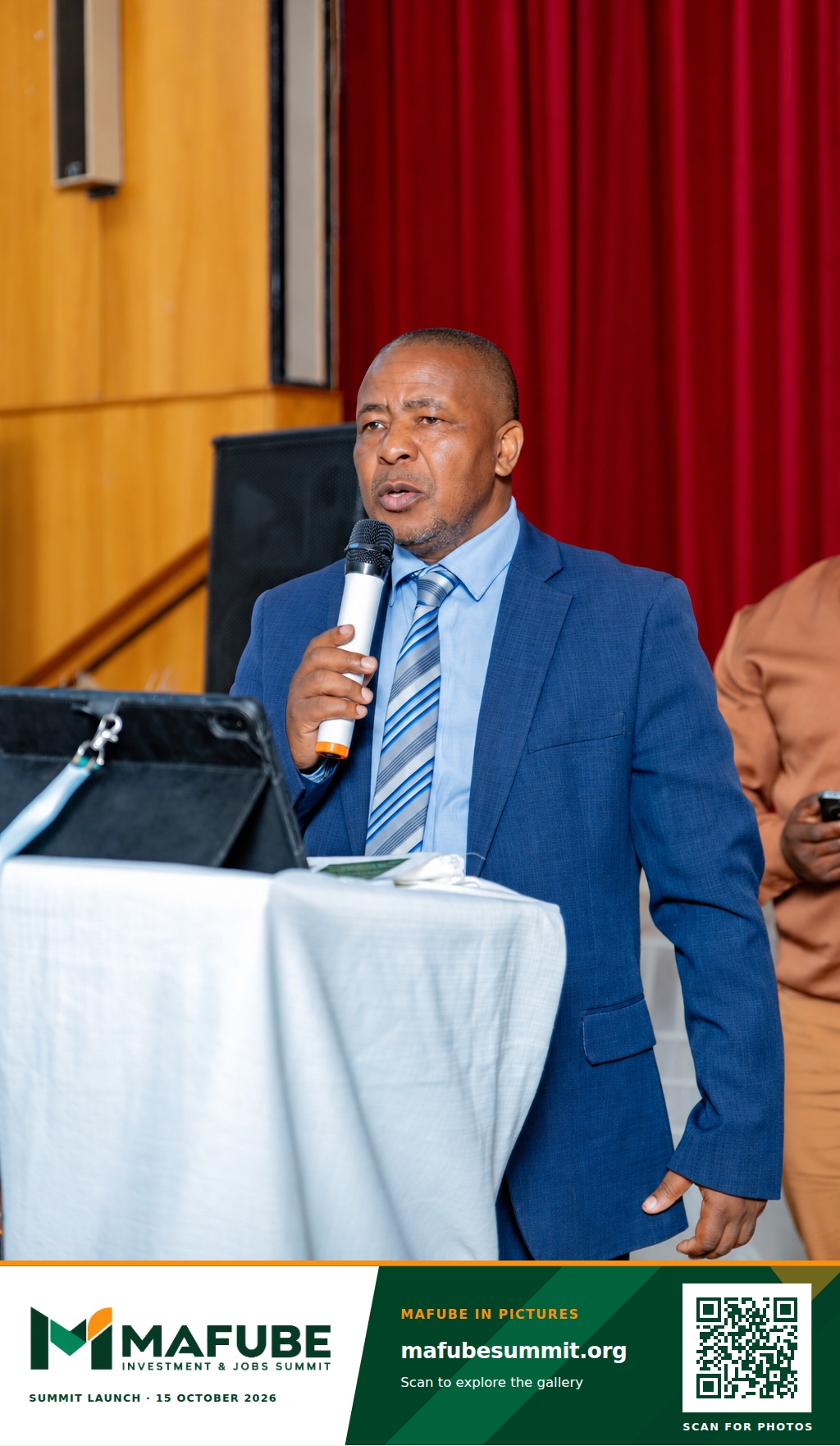 A speaker in a blue suit addresses the hall with a microphone beside the podium (AI-retouched image), with the Mafube Investment & Jobs Summit photo footer